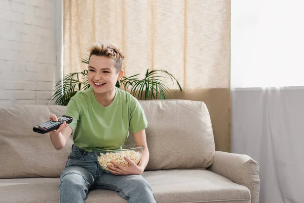 Sonriente persona pensexual con tazón de palomitas de maíz haciendo clic en los canales de televisión en el sofá en casa - foto de stock