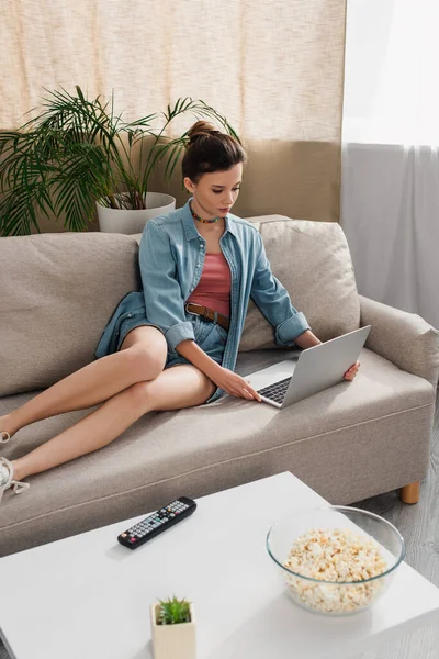 Young woman in denim shirt and jeans using laptop on sofa in living room — Foto stock