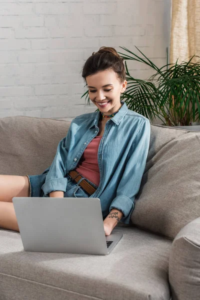 Happy woman in denim shirt using laptop on couch in living room — Stockfoto