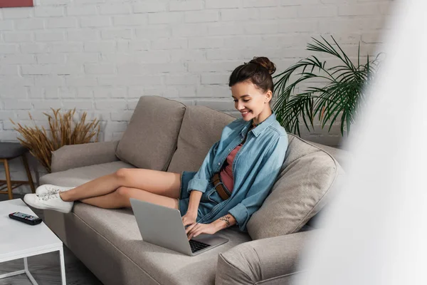 Full length of cheerful woman sitting on couch and typing on laptop on blurred foreground — Fotografia de Stock