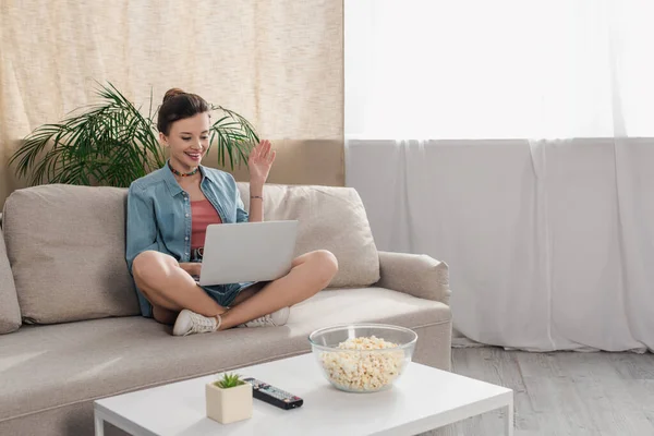 Smiling woman sitting on couch with crossed legs and waving hand during video call on laptop — стоковое фото