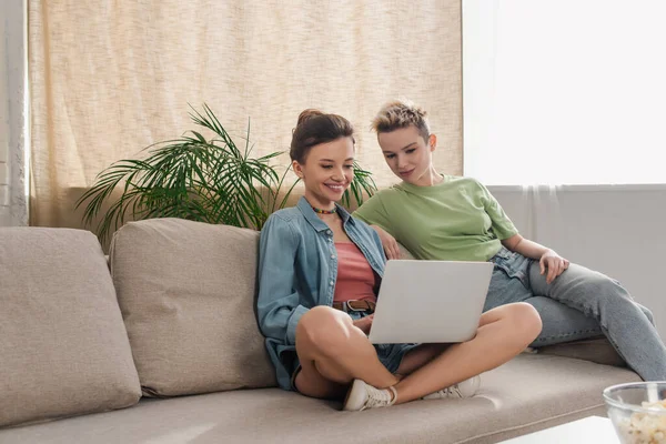 Happy pangender partners watching film on laptop on sofa at home - foto de stock