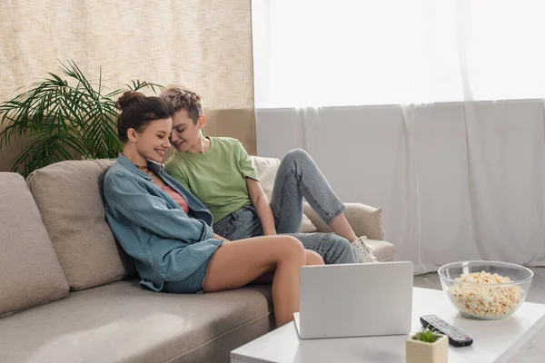 Felici amanti pansessuali seduti sul divano vicino al tavolino con laptop e popcorn — Foto stock