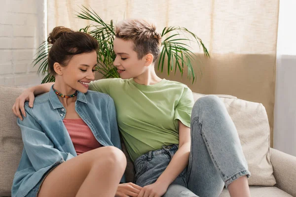 Happy pangender lovers embracing and holding hands on couch at home — Stock Photo