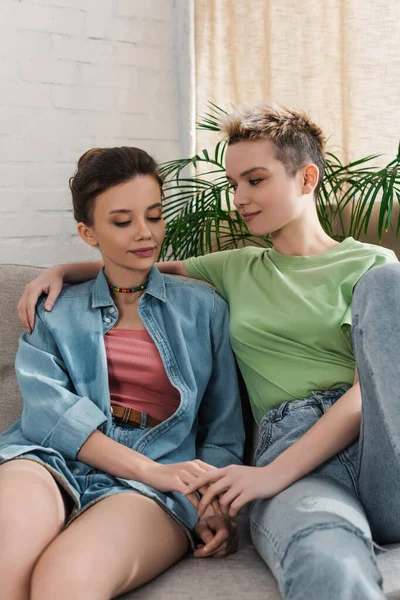 Smiling bigender couple sitting on sofa in living room and holding hands - foto de stock
