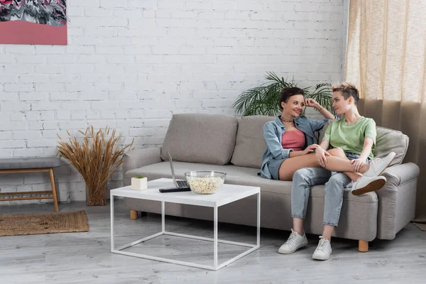 Smiling bigender couple talking on couch in living room near laptop and popcorn on coffee table — Stock Photo