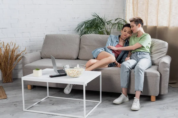 Vista completa de la pareja joven pangender sentado en el sofá cerca de la computadora y palomitas de maíz en la mesa de café - foto de stock