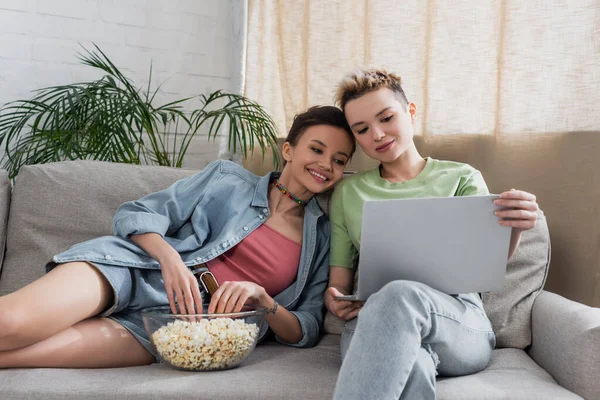 Pleased pansexual couple watching film on laptop near bowl of popcorn on couch — Stock Photo