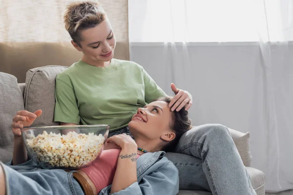 Young bigender partner touching head of happy lover lying on couch with popcorn — стоковое фото