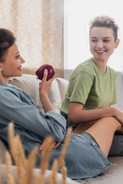 Pleased pansexual person holding ripe apple near young partner at home — стоковое фото
