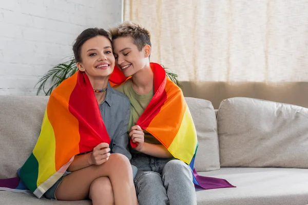 Joyful bigender couple covered with lgbt flag sitting on sofa at home — Stock Photo