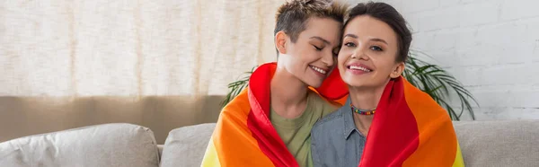 Allegra coppia pansessuale coperta di bandiera lgbt sorridente a casa, banner — Foto stock