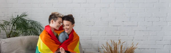 Smiling pangender partners covered with lgbt flag near decorative plants at home, banner — Photo de stock