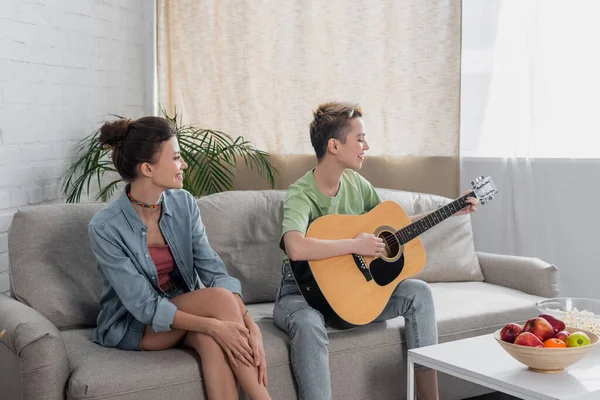Young bigender musician playing acoustic guitar near partner on couch in living room - foto de stock