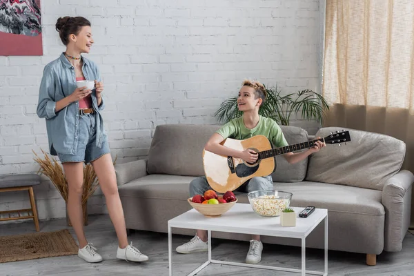 Bigender musician playing guitar on couch near smiling partner standing with cup of tea — Fotografia de Stock