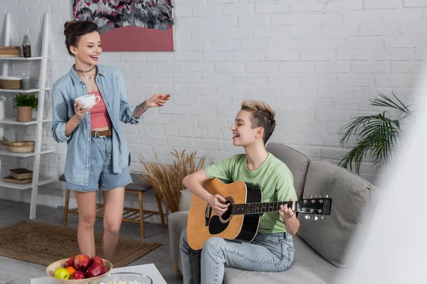 Smiling bigender person with tea cup near partner playing guitar on couch — Foto stock