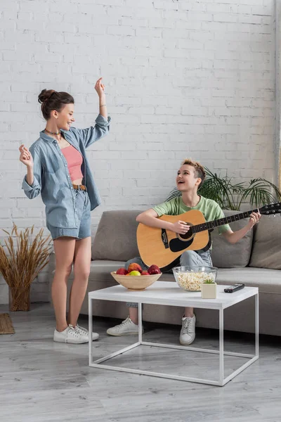 Fröhliche Pangender-Person tanzt neben Musiker, der Gitarre im Wohnzimmer spielt — Stockfoto