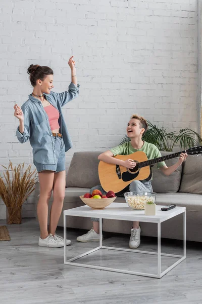 Cheerful pangender person dancing near musician playing guitar in living room — стоковое фото