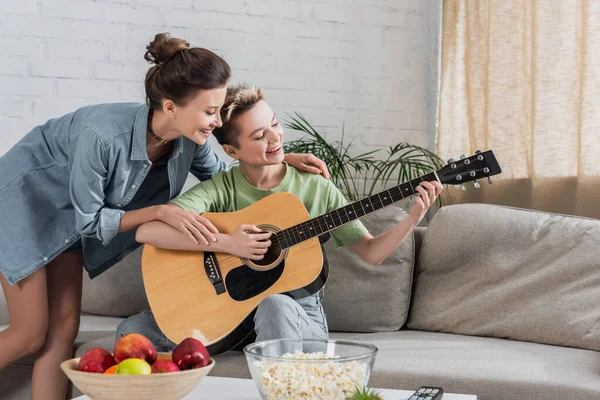 Feliz pangender pessoa tocando guitarra perto do parceiro, tigela de pipoca e maçãs frescas — Fotografia de Stock
