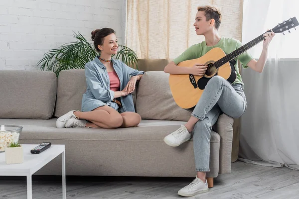 Young pansexual person playing guitar near lover on sofa in living room - foto de stock