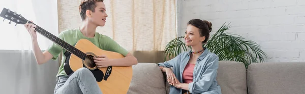 Giovane musicista suonare la chitarra vicino sorridente partner bigender in soggiorno, banner — Foto stock