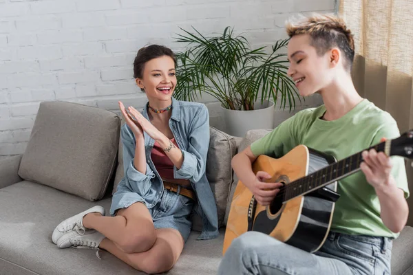 Complacido pangender persona aplaudiendo a amante tocando la guitarra en sala de estar - foto de stock