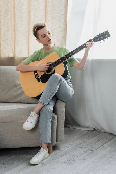 Full length view of young pansexual person in jeans playing acoustic guitar on sofa — стоковое фото