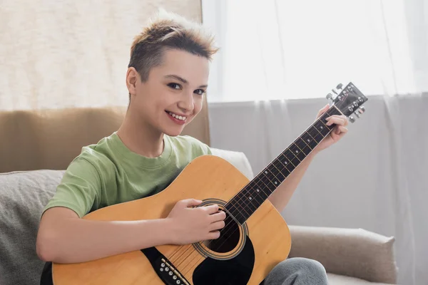 Gai pangender personne regardant la caméra tout en jouant de la guitare acoustique à la maison — Photo de stock