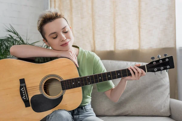 Pleased pansexual person with closed eyes sitting with acoustic guitar in living room — Stockfoto