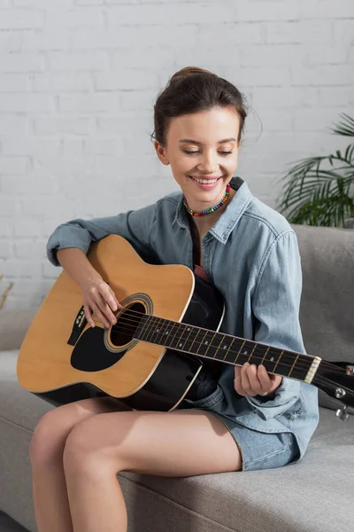 Mujer morena feliz en camisa de mezclilla tocando la guitarra acústica en casa - foto de stock