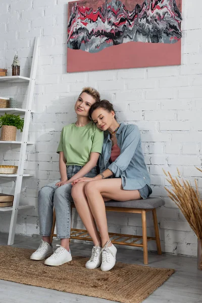 Full length of happy bigender couple sitting under painting near white brick wall — Stock Photo