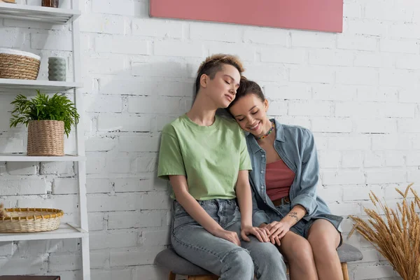 Joven y feliz pangender pareja con los ojos cerrados sentado en otomana cerca de la pared de ladrillo blanco - foto de stock