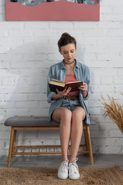 Ansicht der jungen Frau in voller Länge, die in der Nähe weißer Ziegelmauer sitzt und Buch liest — Stockfoto