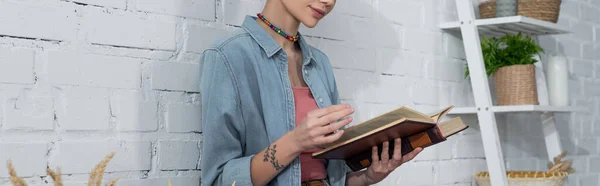 Partial view of tattooed woman in denim shirt reading book near white brick wall, banner — Foto stock