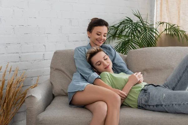 Pleased bigender person embracing partner resting on couch with closed eyes — Stockfoto