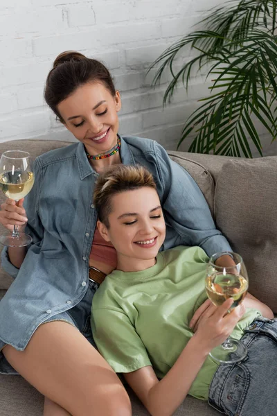 Joyful pansexual couple with glasses of white wine relaxing on sofa at home — Photo de stock