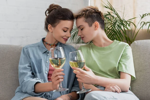 Happy bigender couple sitting with closed eyes and wine glasses at home - foto de stock