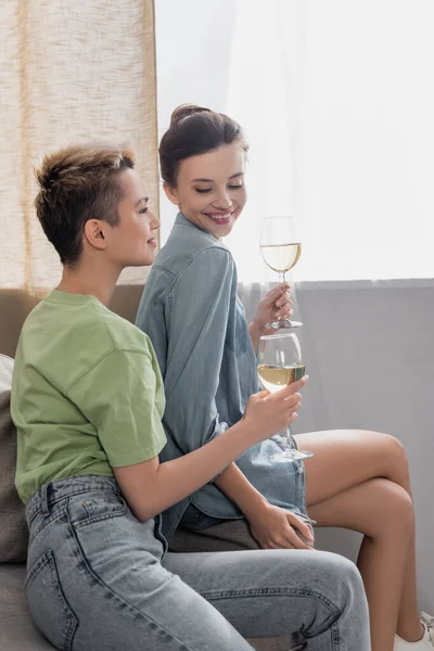 Young pangender couple with glasses of white wine smiling at home - foto de stock