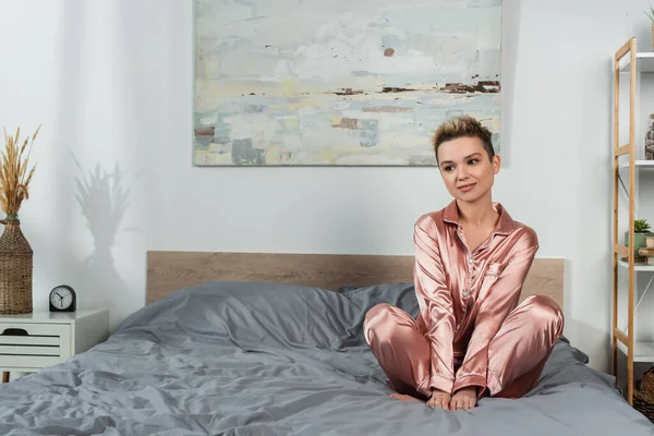 Young and dreamy pangender person in silk pajamas sitting on bed — Stock Photo