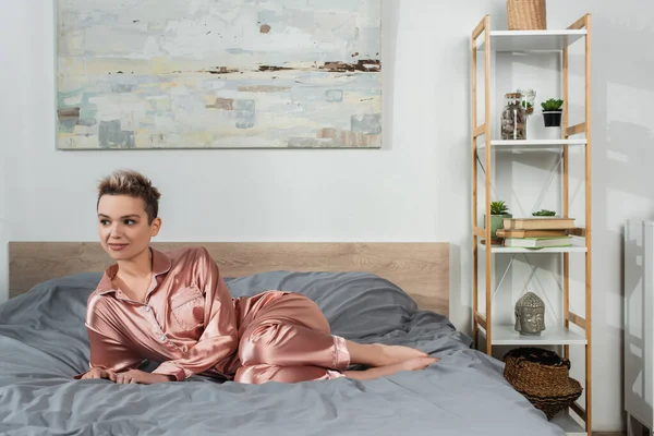 Full length of pangender person in satin pajamas looking away on bed at home — стоковое фото