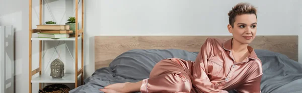 Pretty bigender person in satin pajamas looking away on bed, banner — Photo de stock