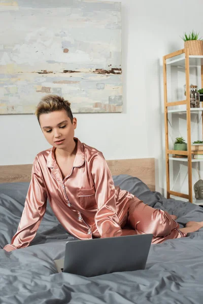 Young pansexual person in silk pajamas using laptop on bed at home — Foto stock