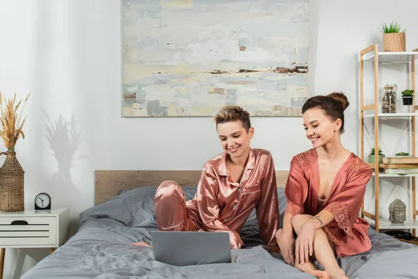 Happy pangender lovers in satin pajamas and robe watching film on laptop in bedroom — Stock Photo