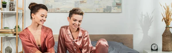 Positive pansexual people in robe and satin pajamas smiling in bedroom, banner - foto de stock