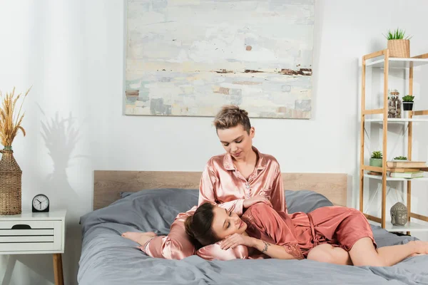 Young pangender person resting on bed near partner in satin pajamas — Photo de stock