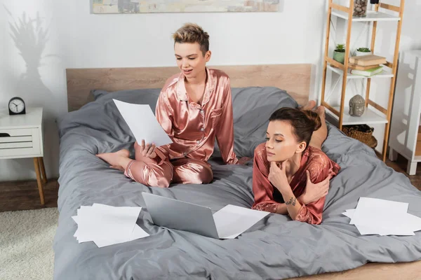 High angle view of young pangender people in sleepwear working with documents and laptop on bed - foto de stock