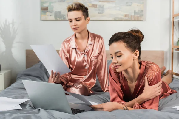 Pansexual people in sleepwear looking at papers while working near laptop on bed — Stock Photo