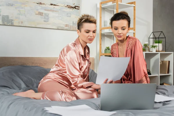 Persone pangender in pigiama di seta e accappatoio che lavorano con carte e laptop sul letto — Foto stock