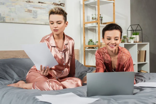 Lächelnde pansexuelle Person arbeitet am Laptop in der Nähe ihres Partners mit Papieren auf dem Bett — Stockfoto