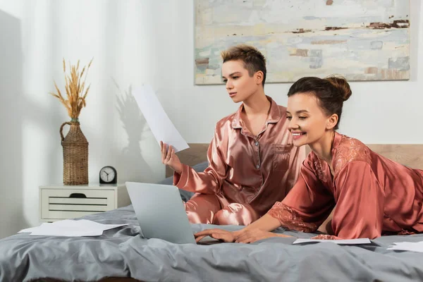 Persona grande feliz usando el ordenador portátil cerca de la pareja que trabaja con papeles en el dormitorio - foto de stock